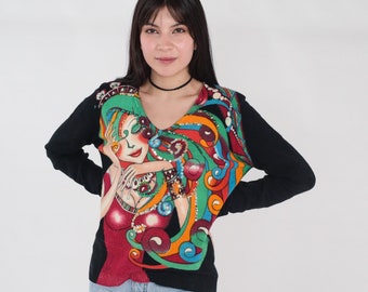 May include: A black long-sleeve sweater featuring a vibrant, multi-colored graphic print of a stylized figure with flowing hair. The design incorporates shades of red, green, orange, and blue, accented with shimmering sequins throughout the artwork.