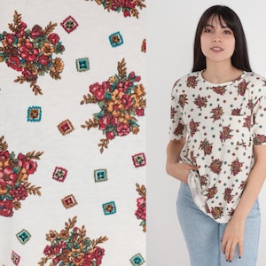 May include: White t-shirt with a floral pattern. The flowers are red, pink, and yellow. The background is white with a subtle texture.
