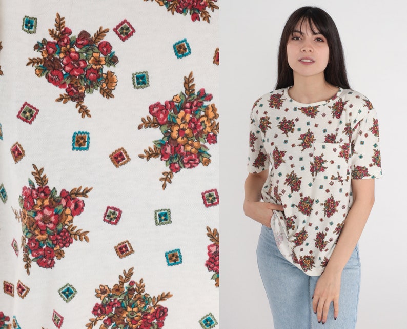 May include: White t-shirt with a floral pattern. The flowers are red, pink, and yellow. The background is white with a subtle texture.