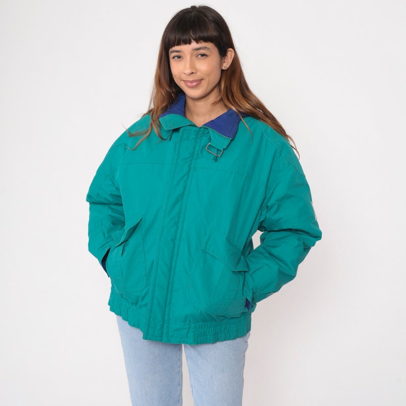 K&ouml;nnte beinhalten: Eine tealblaue Jacke mit einem marineblauen Kragen und einem Rei&szlig;verschluss. Die Jacke hat einen Kordelzug in der Taille und zwei Taschen.