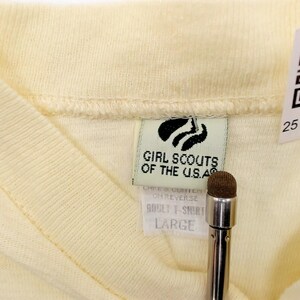 May include: Close-up of a pale yellow V-neck t-shirt. The tag reads "Girl Scouts of the U.S.A." and "ADULT T-SHIRT LARGE". A QR code and a serial number are also visible. The shirt is made of a soft, knit fabric.