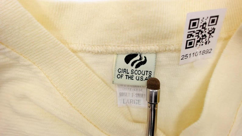 May include: Close-up of a pale yellow V-neck t-shirt. The tag reads "Girl Scouts of the U.S.A." and "ADULT T-SHIRT LARGE". A QR code and a serial number are also visible. The shirt is made of a soft, knit fabric.