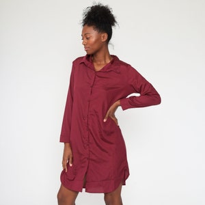 May include: A long-sleeved, button-down shirt dress in a deep burgundy color. The dress features a classic collar and a relaxed fit, with a slightly curved hemline. The fabric appears to have a subtle sheen, suggesting a smooth texture. The dress is knee-length.