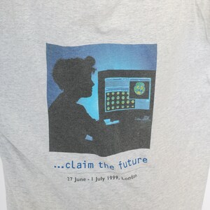 May include: A gray t-shirt with a graphic of a person sitting in front of a computer screen. The text on the shirt reads "...claim the future 27 June - 1 July 1999, London."