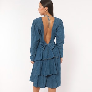 Puede incluir: Un vestido de mezclilla azul de tono medio con mangas largas, espalda descubierta con finos lazos de cierre y un lazo de mezclilla prominente en la cintura. La falda presenta m&uacute;ltiples volantes escalonados que caen en cascada por la espalda.