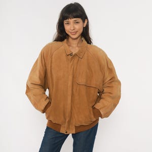 May include: A tan suede jacket with a button closure, a collar, and a brown ribbed hem and cuffs. The jacket has a relaxed fit and a classic design. The model is wearing blue jeans.