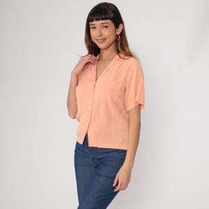 Puede incluir: Una camisa de color melocot&oacute;n de manga corta con cuello y un bolsillo en el pecho. La camisa est&aacute; abotonada por delante y se combina con unos vaqueros azules. La modelo lleva pendientes de aro dorados.