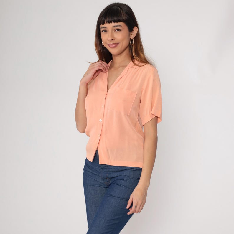 Puede incluir: Una camisa de color melocot&oacute;n de manga corta con cuello y un bolsillo en el pecho. La camisa est&aacute; abotonada por delante y se combina con unos vaqueros azules. La modelo lleva pendientes de aro dorados.