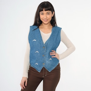 Könnte beinhalten: Eine blaue Jeansweste mit weißen gestickten Delfinen. Die Weste hat einen spitzen Kragen, einen Knopfverschluss und eine figurbetonte Silhouette.