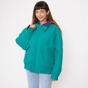 K&ouml;nnte beinhalten: Eine tealblaue Jacke mit einem marineblauen Kragen und einem Rei&szlig;verschluss. Die Jacke hat einen Kordelzug in der Taille und zwei Taschen auf der Vorderseite.