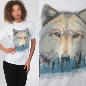 May include: A gray t-shirt with a wolf graphic. The wolf is depicted in a forest setting with blue trees. The wolf's face is the main focus of the graphic.