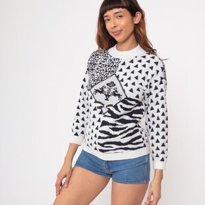 May include: A white and black patterned sweater with a crew neck. The sweater features a mix of geometric and animal print designs, including triangles, zebra stripes, and a diamond-shaped emblem with a crest. The sleeves are three-quarter length.