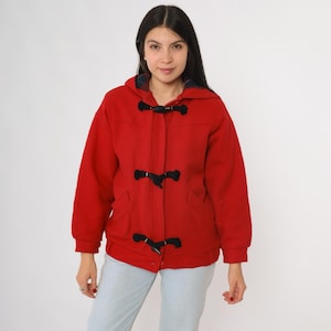 May include: A red hooded duffel coat with black toggle closures. The coat has a classic design with a relaxed fit, featuring a hood and two front pockets. The coat is made of a thick, warm material, perfect for cold weather.