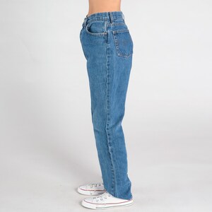 May include: A pair of blue denim jeans with a straight leg fit. The jeans have a high waist and are shown on a model wearing white trainers.