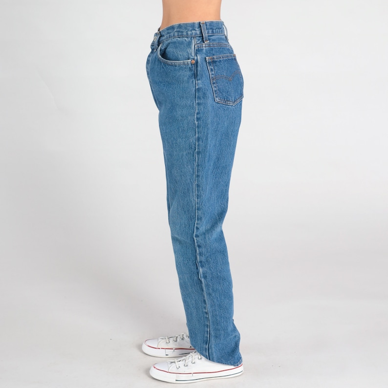 May include: A pair of blue denim jeans with a straight leg fit. The jeans have a high waist and are shown on a model wearing white trainers.
