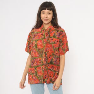 May include: Short-sleeved button-down shirt with a vibrant, abstract pattern in shades of red, green, and purple. The shirt features a collared neckline and a chest pocket. The shirt is made of a lightweight material.