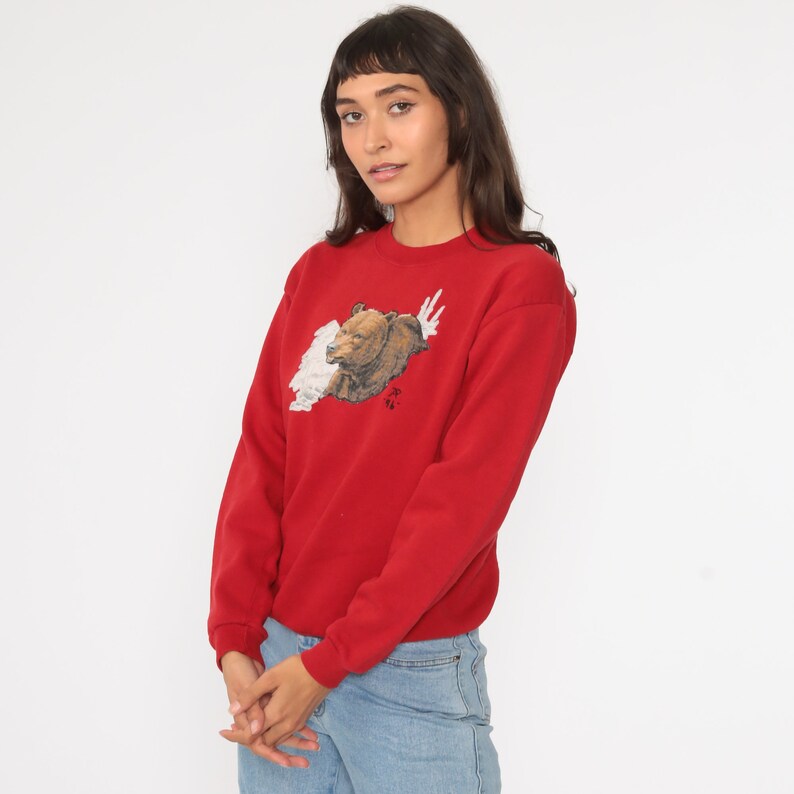 May include: Red sweatshirt with a brown bear graphic on the front. The bear is surrounded by white and has a red symbol on its chest.