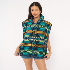 May include: A sleeveless hooded poncho featuring a dark blue base with a vibrant geometric pattern in shades of teal, lime green, yellow, and orange. The garment has a high collar with a partial zipper closure and dark blue trim along the edges.