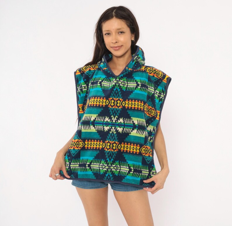 May include: A sleeveless hooded poncho featuring a dark blue base with a vibrant geometric pattern in shades of teal, lime green, yellow, and orange. The garment has a high collar with a partial zipper closure and dark blue trim along the edges.