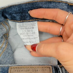 May include: Close-up of blue denim jeans with a white tag. The tag has text including "955BBVW" and "16 REG". The jeans have a brown leather patch with "PREMIUM QUALITY" printed on it. The jeans are made of 100% cotton.