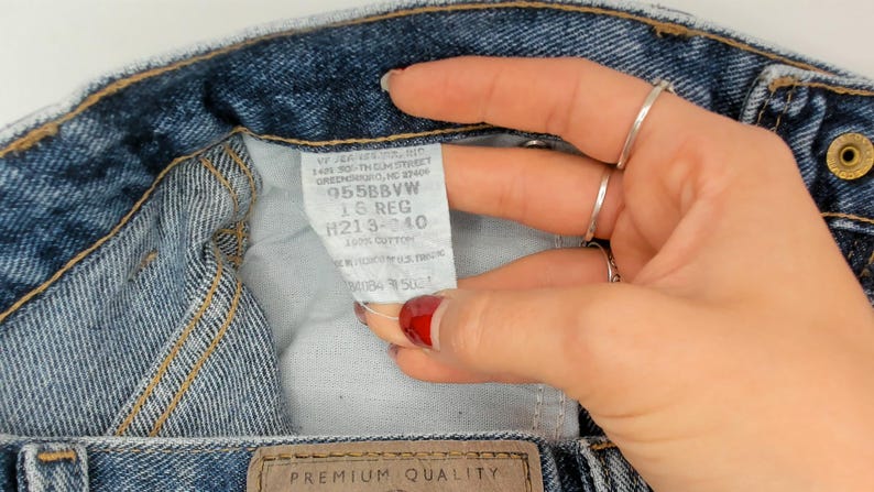 May include: Close-up of blue denim jeans with a white tag. The tag has text including "955BBVW" and "16 REG". The jeans have a brown leather patch with "PREMIUM QUALITY" printed on it. The jeans are made of 100% cotton.