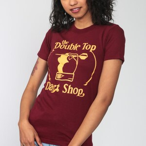 May include: A maroon t-shirt with a gold graphic of a hand throwing a dart at a dartboard. The graphic says "the Double Top Dart Shop".