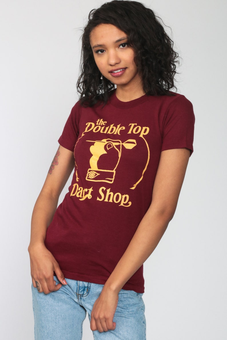 May include: A maroon t-shirt with a gold graphic of a hand throwing a dart at a dartboard. The graphic says "the Double Top Dart Shop".