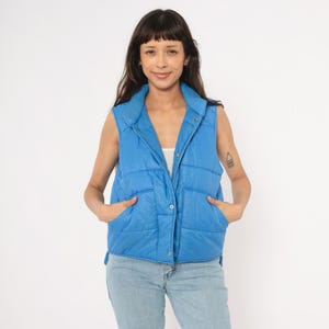 May include: A bright blue, quilted vest with a snap closure and two front pockets. The vest is sleeveless and has a collar. The person is wearing light blue jeans. The vest is a vibrant, solid color.