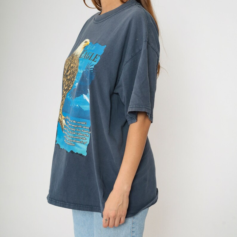 May include: A navy blue t-shirt featuring a graphic of a bald eagle with the text "Bald Eagle" on the front. The eagle is perched on a branch with a blue and white background. The text below the eagle reads "One of the world's most majestic birds, the only member of the genus Haliaeetus native to North America. The presence of the bald eagle is a testament to the health of the ecosystem."