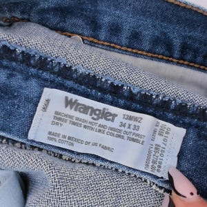 May include: Close-up of blue denim Wrangler jeans. The label reads "Wrangler 13MWZ 34 X 33". The jeans are made of 100% cotton and made in Mexico of US fabric. The care instructions are also visible on the label.