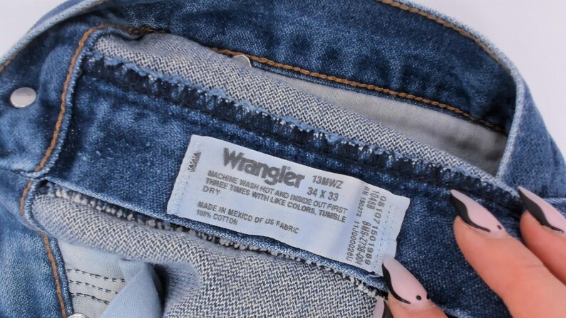 May include: Close-up of blue denim Wrangler jeans. The label reads "Wrangler 13MWZ 34 X 33". The jeans are made of 100% cotton and made in Mexico of US fabric. The care instructions are also visible on the label.