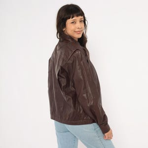 May include: A dark brown leather jacket with a classic bomber style. The jacket features a zip-up front, shoulder pads, and a ribbed hem. The model is wearing light blue jeans.