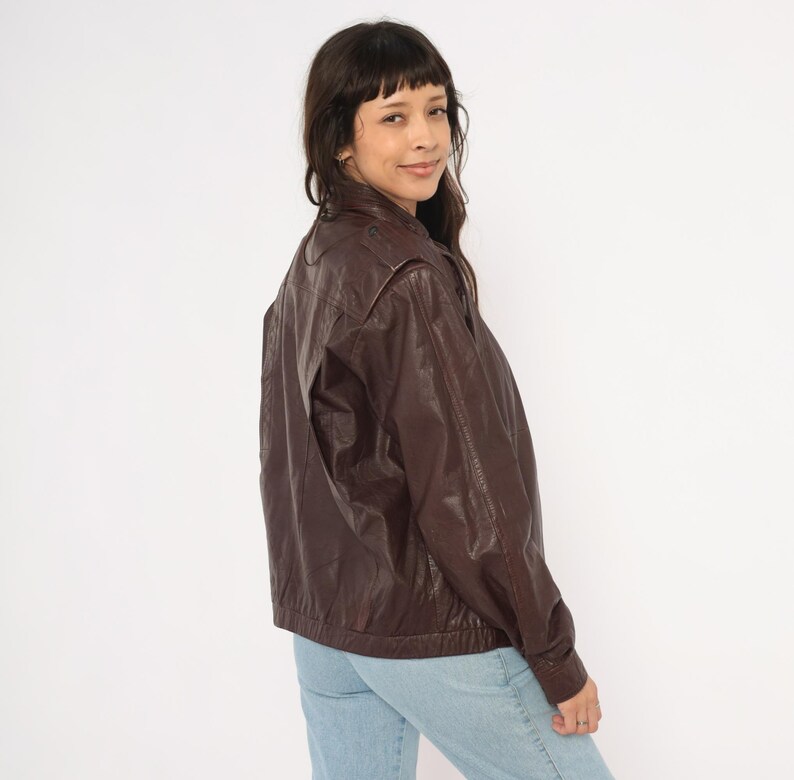 May include: A dark brown leather jacket with a classic bomber style. The jacket features a zip-up front, shoulder pads, and a ribbed hem. The model is wearing light blue jeans.