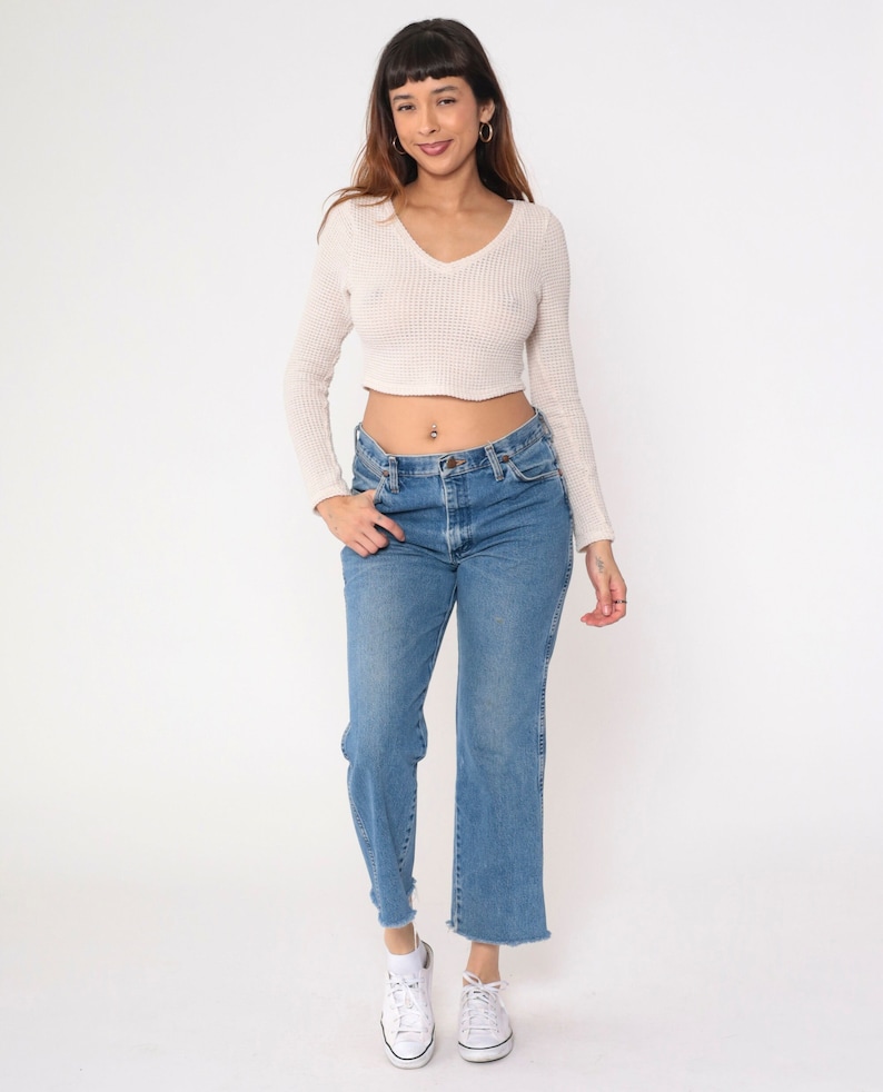 May include: A woman wearing a white cropped waffle knit sweater with long sleeves and a v-neck. She is wearing blue denim jeans with a frayed hem and white sneakers.