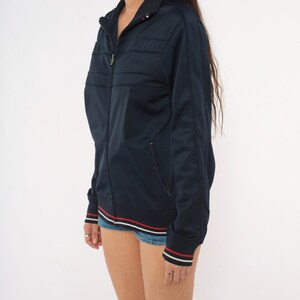 May include: A dark navy blue zip-up track jacket featuring a high collar, long sleeves, and two front pockets. The hem and sleeve cuffs are detailed with thin white and red horizontal stripes. The jacket has a smooth, synthetic fabric finish.