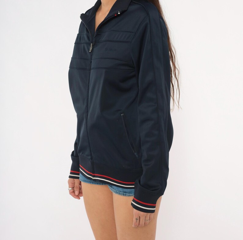 May include: A dark navy blue zip-up track jacket featuring a high collar, long sleeves, and two front pockets. The hem and sleeve cuffs are detailed with thin white and red horizontal stripes. The jacket has a smooth, synthetic fabric finish.