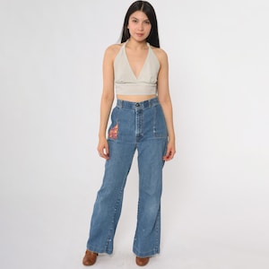 May include: A woman wearing a beige halter top with a striped pattern and high-waisted, light-wash blue jeans. The jeans have a decorative patch on the front pocket and a wide leg. The woman is wearing brown shoes.