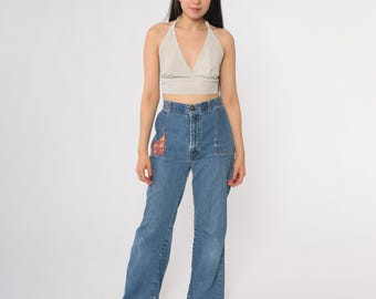 May include: A woman wearing a beige halter top with a striped pattern and high-waisted, light-wash blue jeans. The jeans have a decorative patch on the front pocket and a wide leg. The woman is wearing brown shoes.