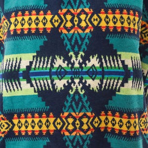 May include: A textile fabric featuring a repeating geometric pattern in dark blue, teal, yellow, orange, and white. The design consists of horizontal bands and symmetrical diamond-shaped motifs with intricate jagged edges and linear details.
