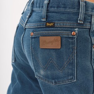 Peut inclure: Gros plan sur un jean en denim bleu avec un patch en cuir marron "Wrangler" sur la poche arri&egrave;re. Le jean a des rivets en cuivre et une petite &eacute;tiquette "Wrangler" au-dessus de la poche arri&egrave;re. Un style de jean classique.