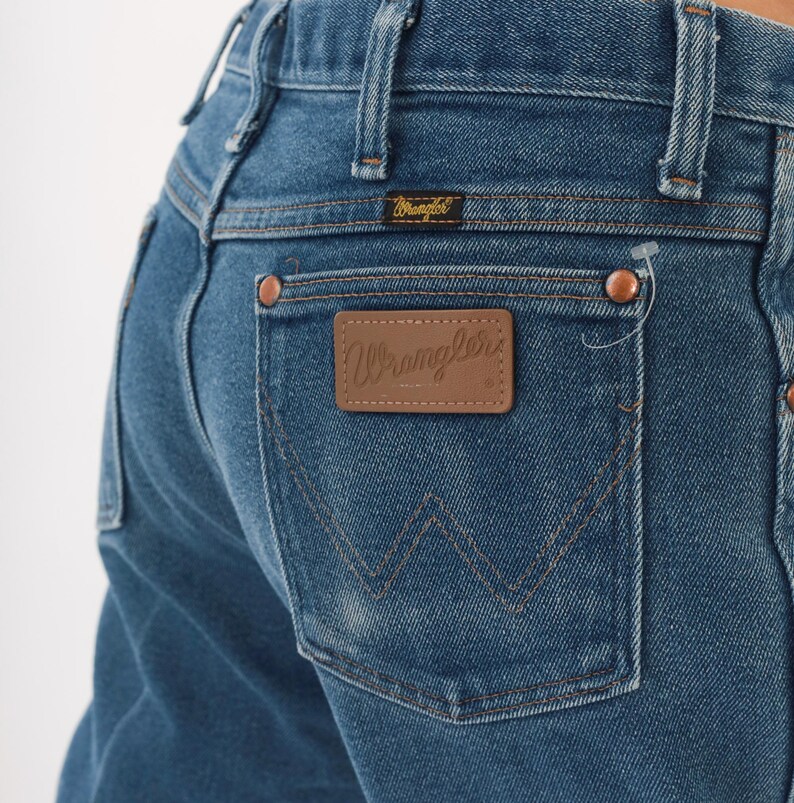 Peut inclure: Gros plan sur un jean en denim bleu avec un patch en cuir marron "Wrangler" sur la poche arri&egrave;re. Le jean a des rivets en cuivre et une petite &eacute;tiquette "Wrangler" au-dessus de la poche arri&egrave;re. Un style de jean classique.