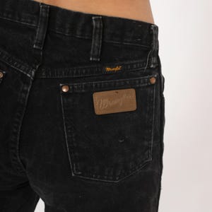 May include: Close-up of black denim jeans. The jeans have a classic five-pocket design with copper rivets and a brown leather patch on the back pocket with the brand name "Wrangler". The jeans are made of a durable denim fabric.