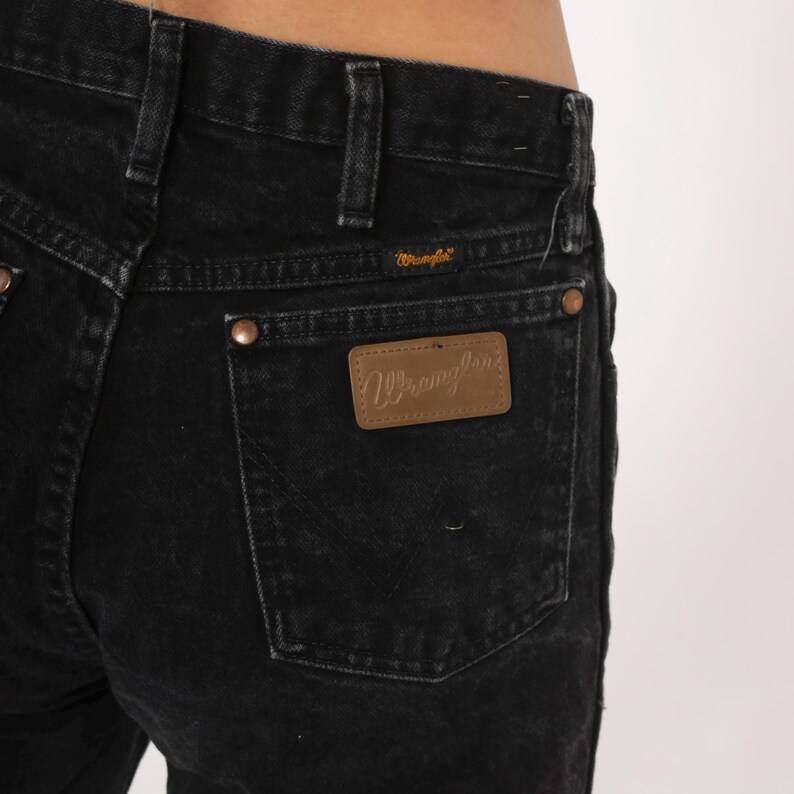 May include: Close-up of black denim jeans. The jeans have a classic five-pocket design with copper rivets and a brown leather patch on the back pocket with the brand name "Wrangler". The jeans are made of a durable denim fabric.