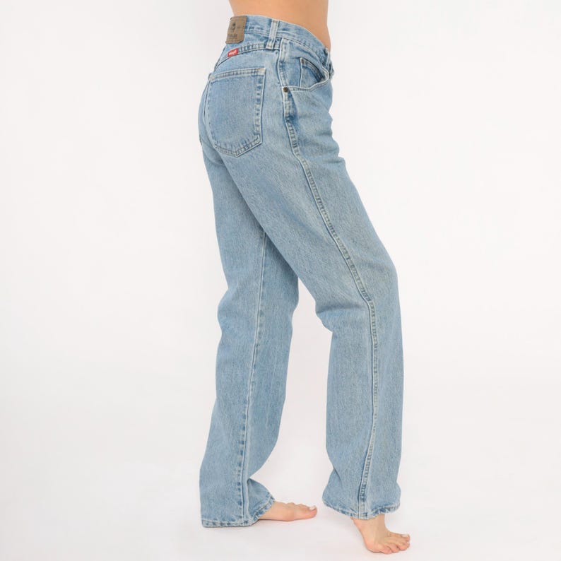 May include: Light blue denim jeans with a classic straight leg fit. The jeans feature a high waist, belt loops, and a small leather patch on the back. The jeans are a light wash and have a faded appearance.