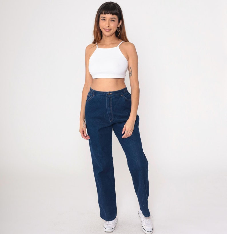K&ouml;nnte beinhalten: Ein wei&szlig;es Neckholder-Top und dunkelblaue Jeans. Die Jeans haben eine hohe Taille und eine gerade Beinpassform. Das Model tr&auml;gt wei&szlig;e Turnschuhe.