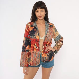 May include: A long-sleeved open-front jacket featuring a vibrant batik-style pattern in shades of red, orange, blue, and black. The garment has a straight hem, a simple collarless neckline, and a relaxed fit, showcasing intricate floral and geometric motifs.