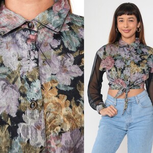 May include: A floral print shirt with a collar and sheer black sleeves. The shirt is tied at the waist. The floral pattern incorporates shades of purple, green, and gold.