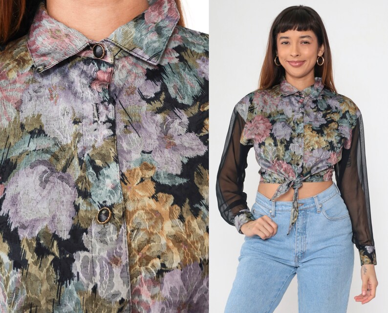 May include: A floral print shirt with a collar and sheer black sleeves. The shirt is tied at the waist. The floral pattern incorporates shades of purple, green, and gold.