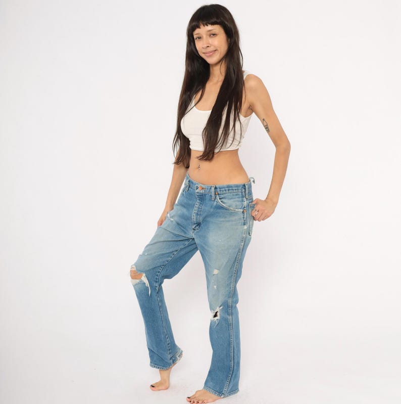 May include: Light blue denim jeans with distressed details, including ripped knees. The jeans have a classic five-pocket design and a relaxed fit. The model is wearing a white crop top.