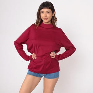 Puede incluir: Una camiseta burdeos de manga larga con cuello alto. La camiseta tiene un corte holgado y se combina con pantalones cortos de mezclilla. La modelo está de pie frente a un fondo blanco.